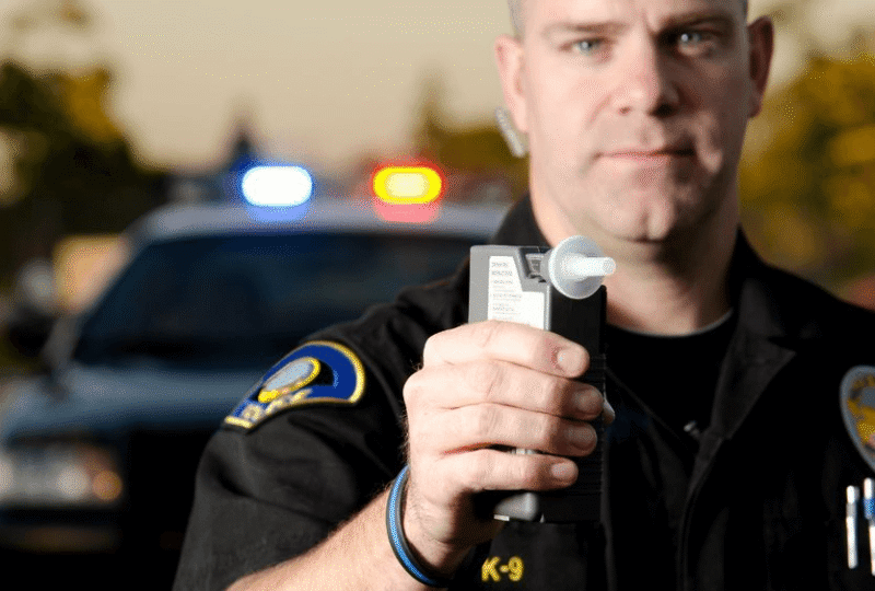 Police officer holding a breathalyzer in front of a patrol car, symbolizing Vermont DUI enforcement and license reinstatement processes relevant to auto insurance.