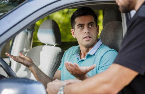 A police officer has pulled over a male driver in a blue shirt, who makes a gesture with his hands indicating he does not have proof of valid Colorado auto liability coverage due to a payment lapse.
