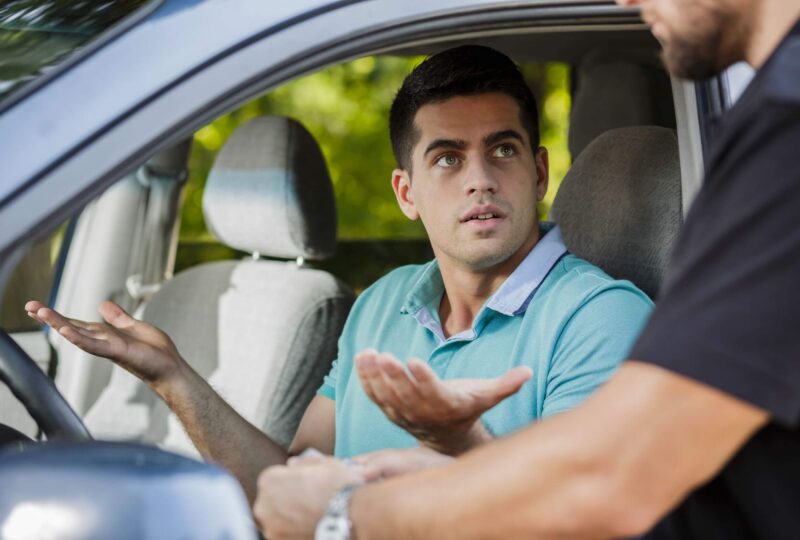 A police officer has pulled over a male driver in a blue shirt, who makes a gesture with his hands indicating he does not have proof of valid Colorado auto liability coverage due to a payment lapse.