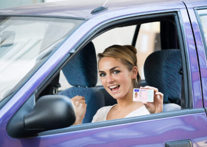A happy young woman sitting behind the wheel of a purple car and displaying her driver's license after following Colorado license reintatement requirements to restore her driving privileges.