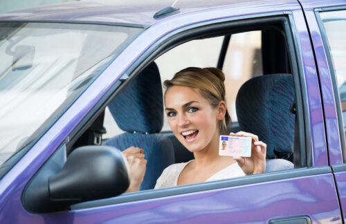 A happy young woman sitting behind the wheel of a purple car and displaying her driver's license after following Colorado license reintatement requirements to restore her driving privileges.