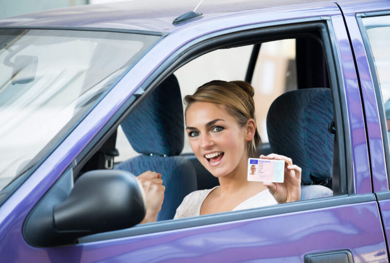 A happy young woman sitting behind the wheel of a purple car and displaying her driver's license after following Colorado license reintatement requirements to restore her driving privileges.