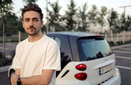 Young man in a white t-shirt standing in front of a small white car after learning how long he will need SR22 insurance in Colorado.