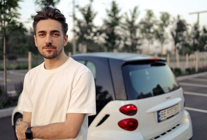 Young man in a white t-shirt standing in front of a small white car after learning how long he will need SR22 insurance in Colorado.