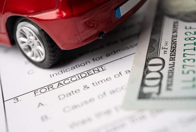 A toy car, an accident report, and a one-hundred dollar bill signifying Colorado SR22 insurance costs.