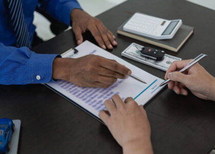 An insurance agent helps a customer compare non-owner SR22 quotes. An insurance document, car keys, a calculator, and cash on the desk indicate their discussion of policy rates.