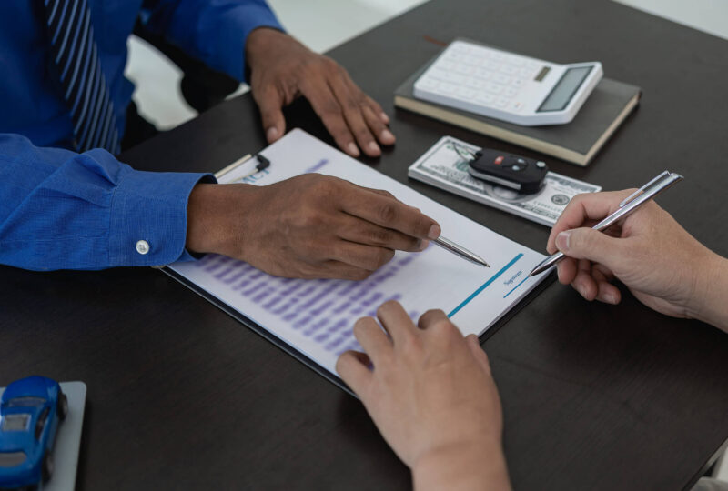 An insurance agent helps a customer compare non-owner SR22 quotes. An insurance document, car keys, a calculator, and cash on the desk indicate their discussion of policy rates.
