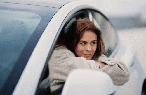 A young woman looks out of the driver's seat window of a white car with an expression conveying uncertainty about how long she needs to maintain SR22 insurance in Kansas.