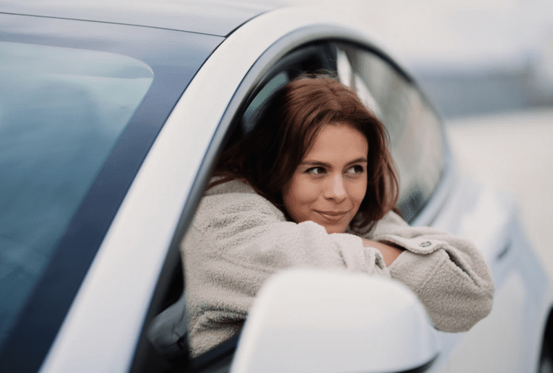 A young woman looks out of the driver's seat window of a white car with an expression conveying uncertainty about how long she needs to maintain SR22 insurance in Kansas.