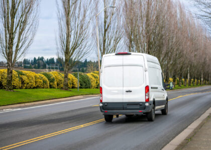 A white commercial delivery van on a road lined with trees and yellow shrubs presents a question: If a driver personally needs SR-22 insurance, can they get commercial SR-22 insurance?