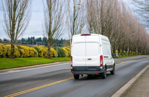 A white commercial delivery van on a road lined with trees and yellow shrubs presents a question: If a driver personally needs SR-22 insurance, can they get commercial SR-22 insurance?