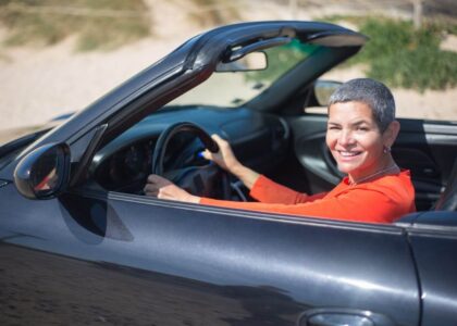 A smiling, middle-aged woman in a red shirt driving a black convertible who has reinstated her driver&rsquo;s license after her Alabama SR22 insurance lapsed.