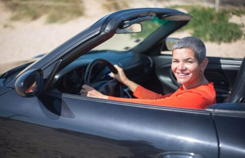 A smiling, middle-aged woman in a red shirt driving a black convertible who has reinstated her driver’s license after her Alabama SR22 insurance lapsed.