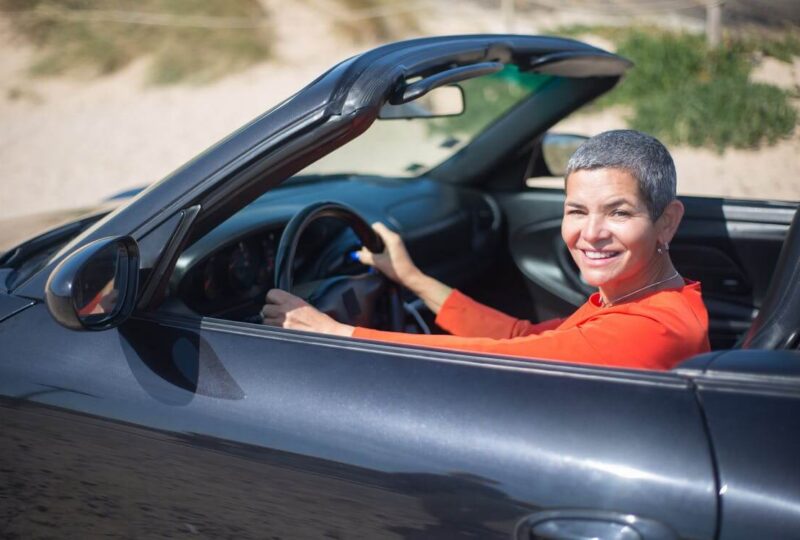 A smiling, middle-aged woman in a red shirt driving a black convertible who has reinstated her driver&rsquo;s license after her Alabama SR22 insurance lapsed.