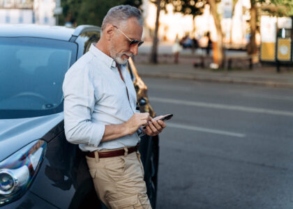 A middle-aged man in a blue shirt & khakis leans against the side of his car, using his cell phone to research what to do if his Kansas SR22 policy lapses.