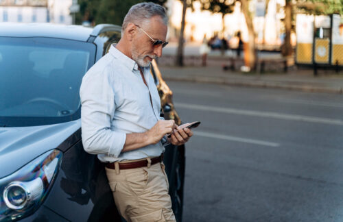 A middle-aged man in a blue shirt & khakis leans against the side of his car, using his cell phone to research what to do if his Kansas SR22 policy lapses.