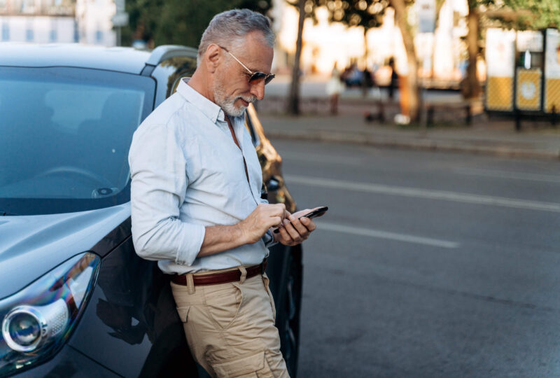 A middle-aged man in a blue shirt & khakis leans against the side of his car, using his cell phone to research what to do if his Kansas SR22 policy lapses.