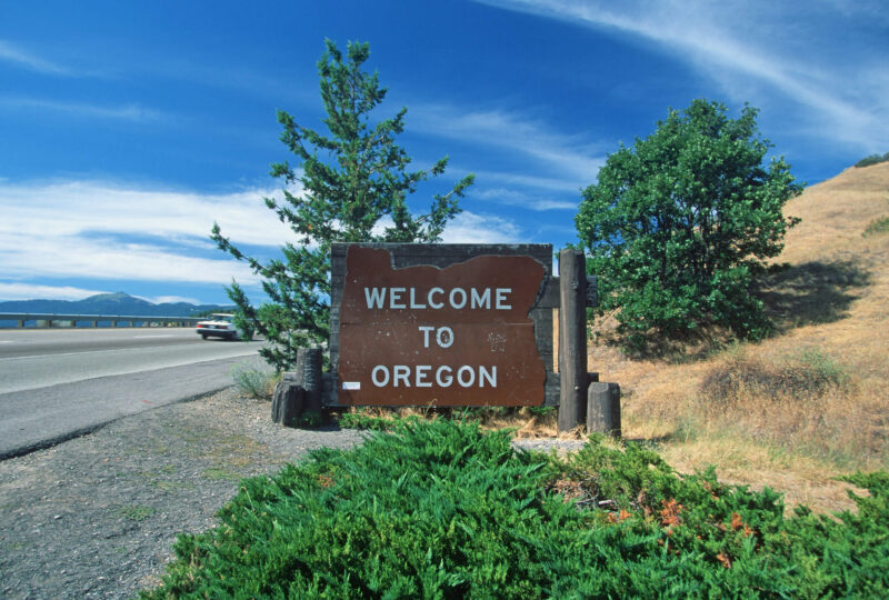 A Welcome to Oregon highway sign on a scenic coastal highway, representing the state's requirement for the length of time drivers need to maintain SR22 insurance.