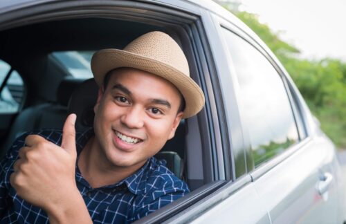 A smiling man in a hat sits behind the wheel of a car giving thumbs up after obtaining an Oregon non-owner (operator) SR22 insurance filing for license reinstatement.