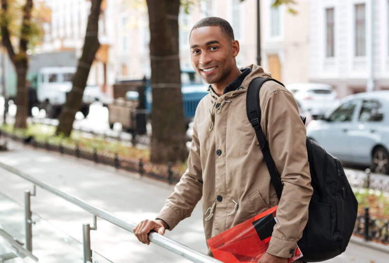 A young man pausing at a sidewalk railing carrying a backpack and red folder, reflecting on how long he will need to keep SR22 insurance in Tennessee.